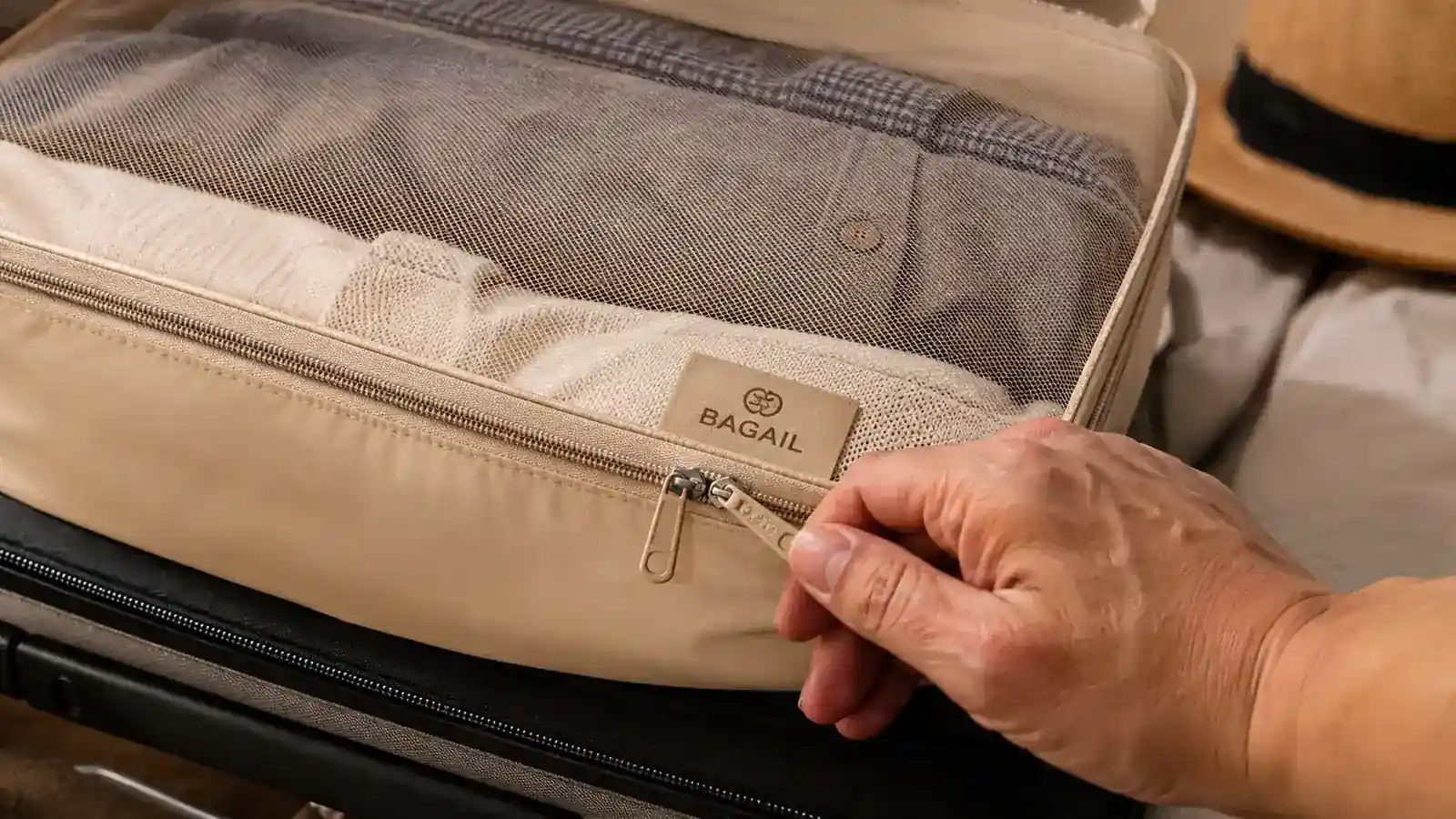 A hand zipping closed a BAGAIL packing cube filled with folded t-shirts, showing the mesh top panel
