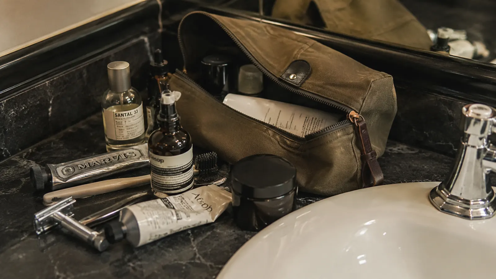 A dopp kit fully unpacked and spread across a small hotel bathroom counter with items sliding toward the sink