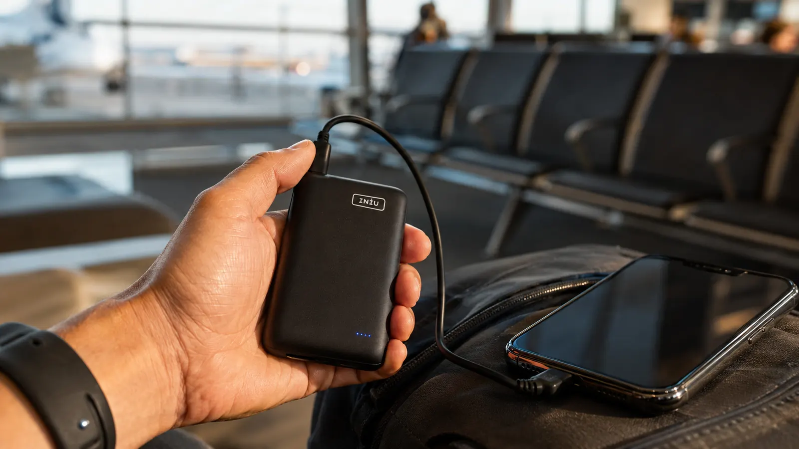 A hand holding the INIU power bank with its detachable USB-C cable plugged into a smartphone at a departure gate