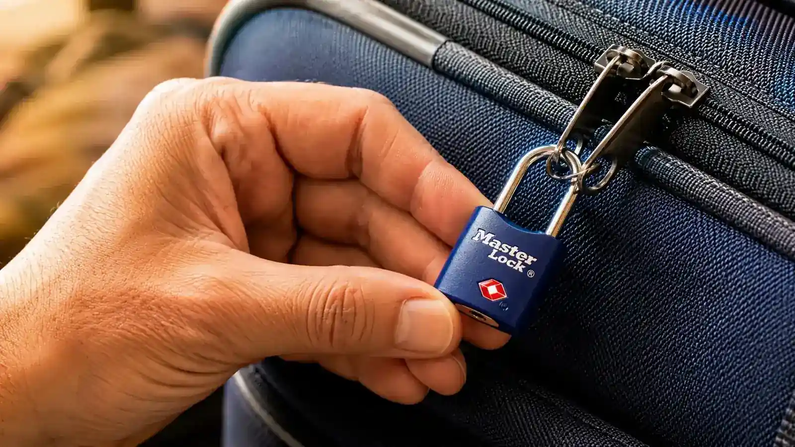 A hand threading the Master Lock 4689T keyed shackle through two zipper pulls on a suitcase