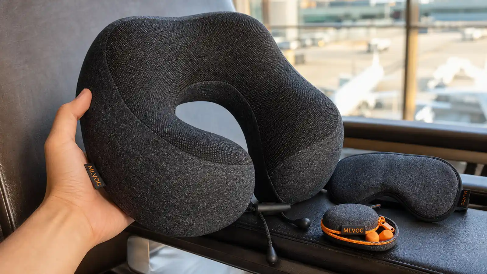 Hand holding a curved gray MLVOC memory foam neck pillow next to a folded eye mask and a small earplug case