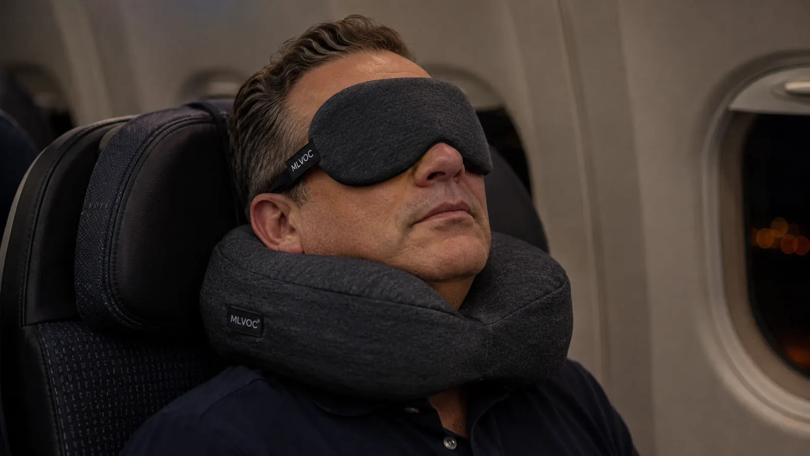 Traveler asleep upright in an airplane seat wearing a curved memory foam neck pillow and eye mask, cabin dark